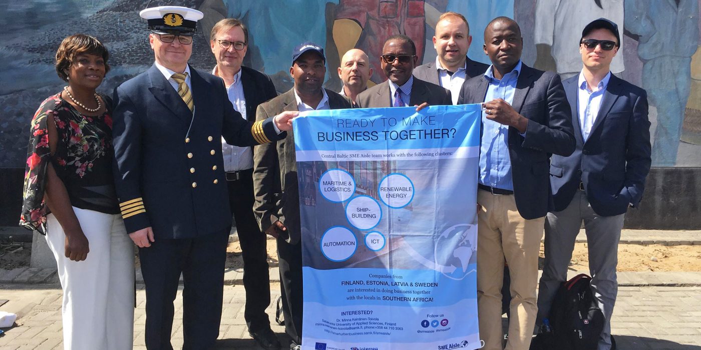 Representatives of companies from the Central Baltic Region in a group photo representatives of Namport organization outdoors in Walvis Bay. Two persons holding a textile flag of the core themes of the SME Aisle project.