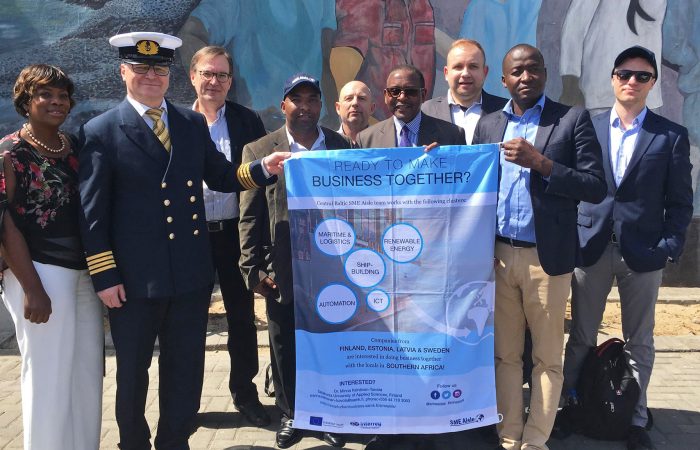 Representatives of companies from the Central Baltic Region in a group photo representatives of Namport organization outdoors in Walvis Bay. Two persons holding a textile flag of the core themes of the SME Aisle project.