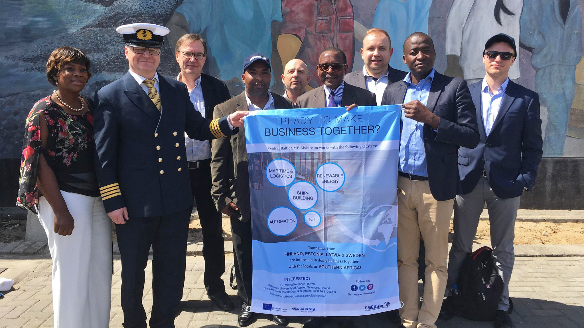 Representatives of companies from the Central Baltic Region in a group photo representatives of Namport organization outdoors in Walvis Bay. Two persons holding a textile flag of the core themes of the SME Aisle project.