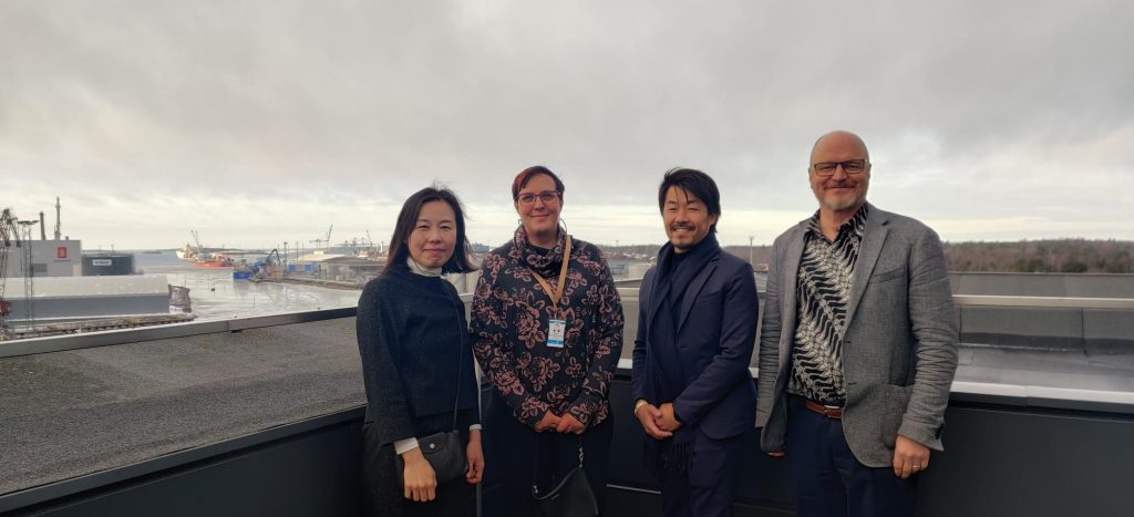 Yusuke Mori, Minna Keinänen-Toivola, Shigemi Matsuzaki and Heikki Koivisto stand in a row on the observation deck of SAMK's Rauma campus. Rauma harbour is visible in the background.