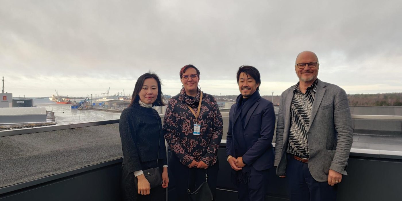 Yusuke Mori, Minna Keinänen-Toivola, Shigemi Matsuzaki and Heikki Koivisto stand in a row on the observation deck of SAMK's Rauma campus. Rauma harbour is visible in the background.