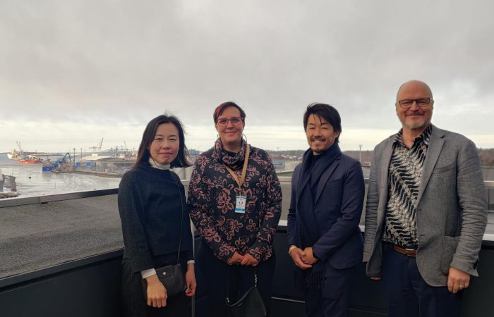 Yusuke Mori, Minna Keinänen-Toivola, Shigemi Matsuzaki and Heikki Koivisto stand in a row on the observation deck of SAMK's Rauma campus. Rauma harbour is visible in the background.