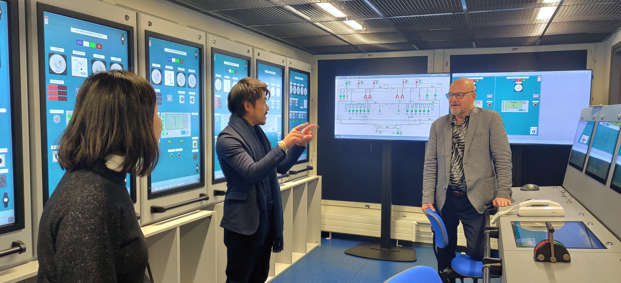 Yusuke Mori, Shigemi Matsuzaki ja Heikki Koivisto keskustelevat simulaatiotilojen kontrollihuoneessa. Taustalla isoja näyttöjä, joissa kaaviokuvia.