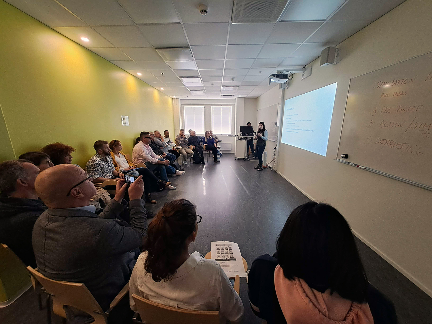 Julia Lepistö is presenting her thesis. There is a PowerPoint presentation in the background. The audience is sitting in chairs, listening.