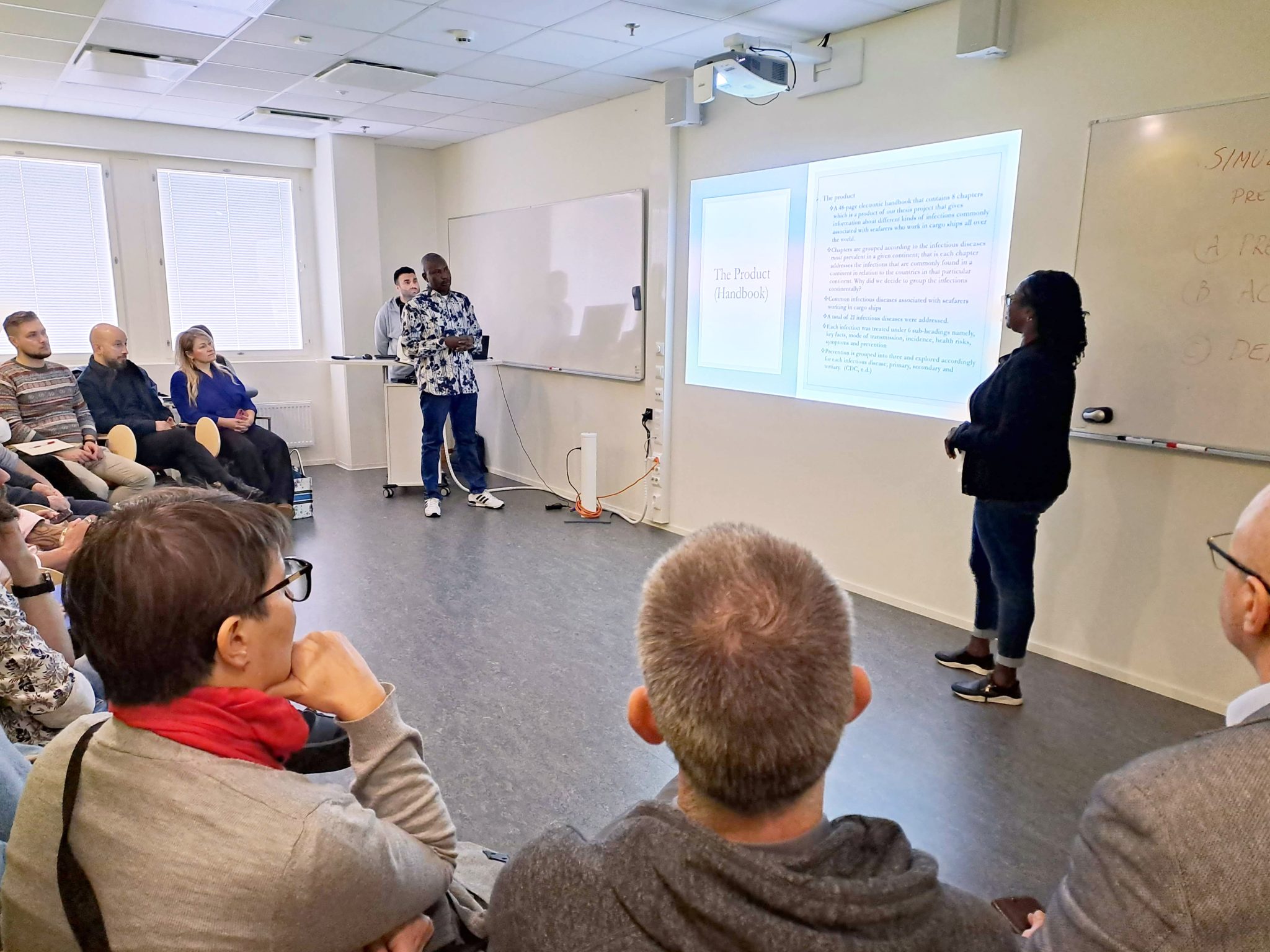 Dessev Vierailu3 Ünal Selim (left), Ajayi Adeniyi, and Janet Anomwa Pitkänen stand in front of the class with a PowerPoint presentation in the background. The audience is sitting in chairs, listening.