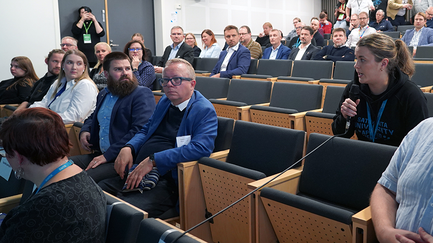 Tutkimusfoorumin yleisöä auditoriossa. Yleisöstä yksi henkilö puhuu mikrofoniin.