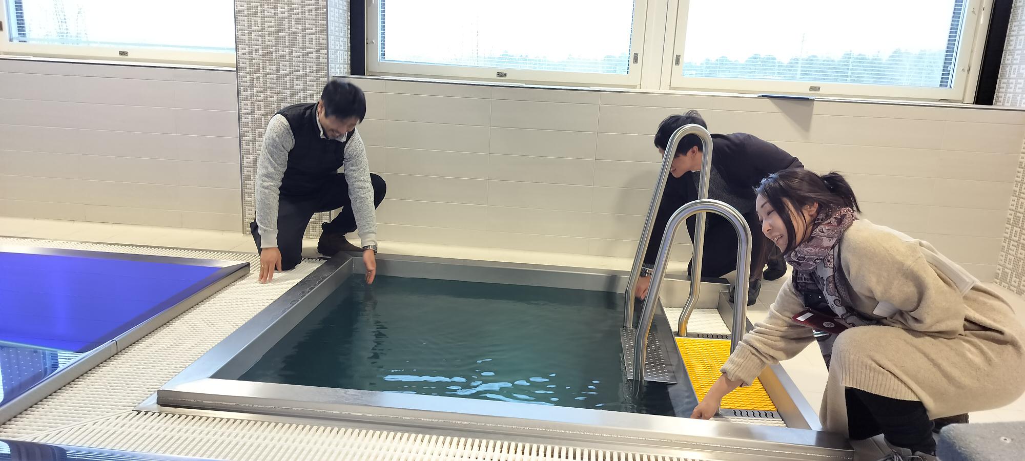 Three people crouching down to test the water temperature of a small cold plunge pool.