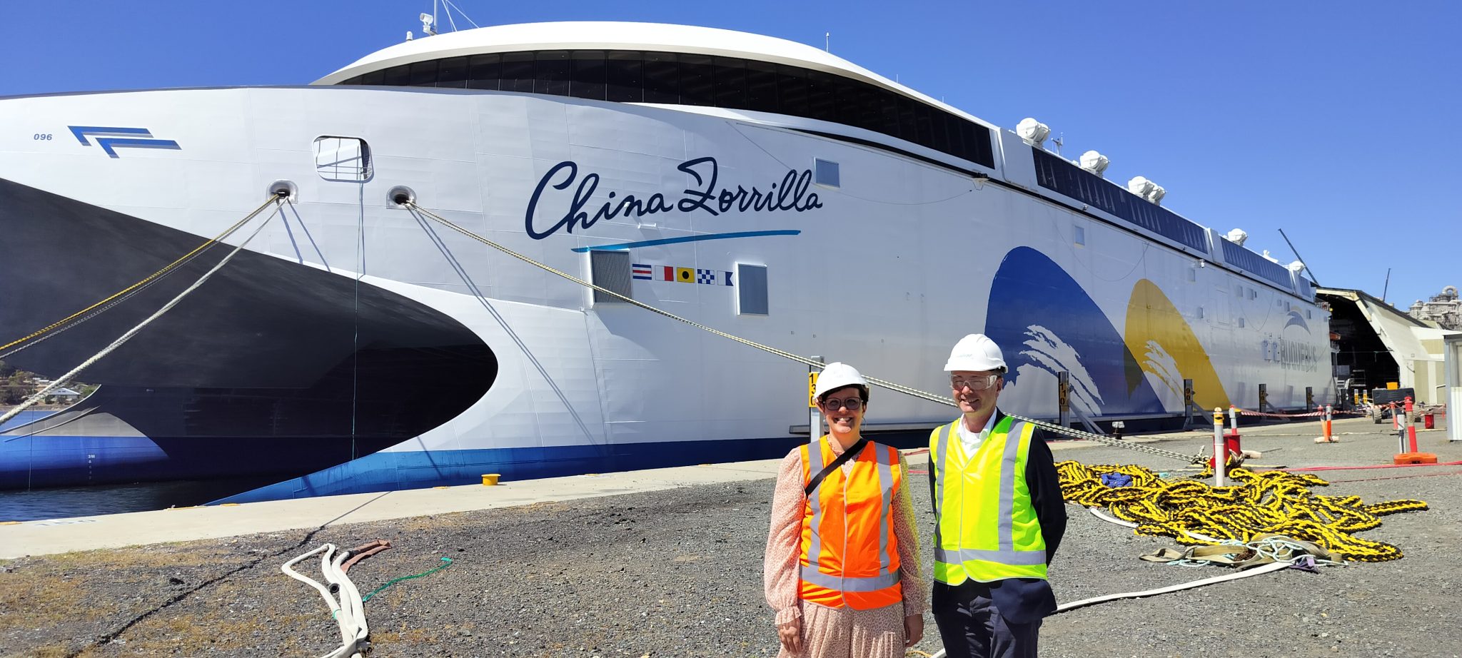 Two people at a port, wearing safety helmets and high-visibility vests. A ship is visible in the background.