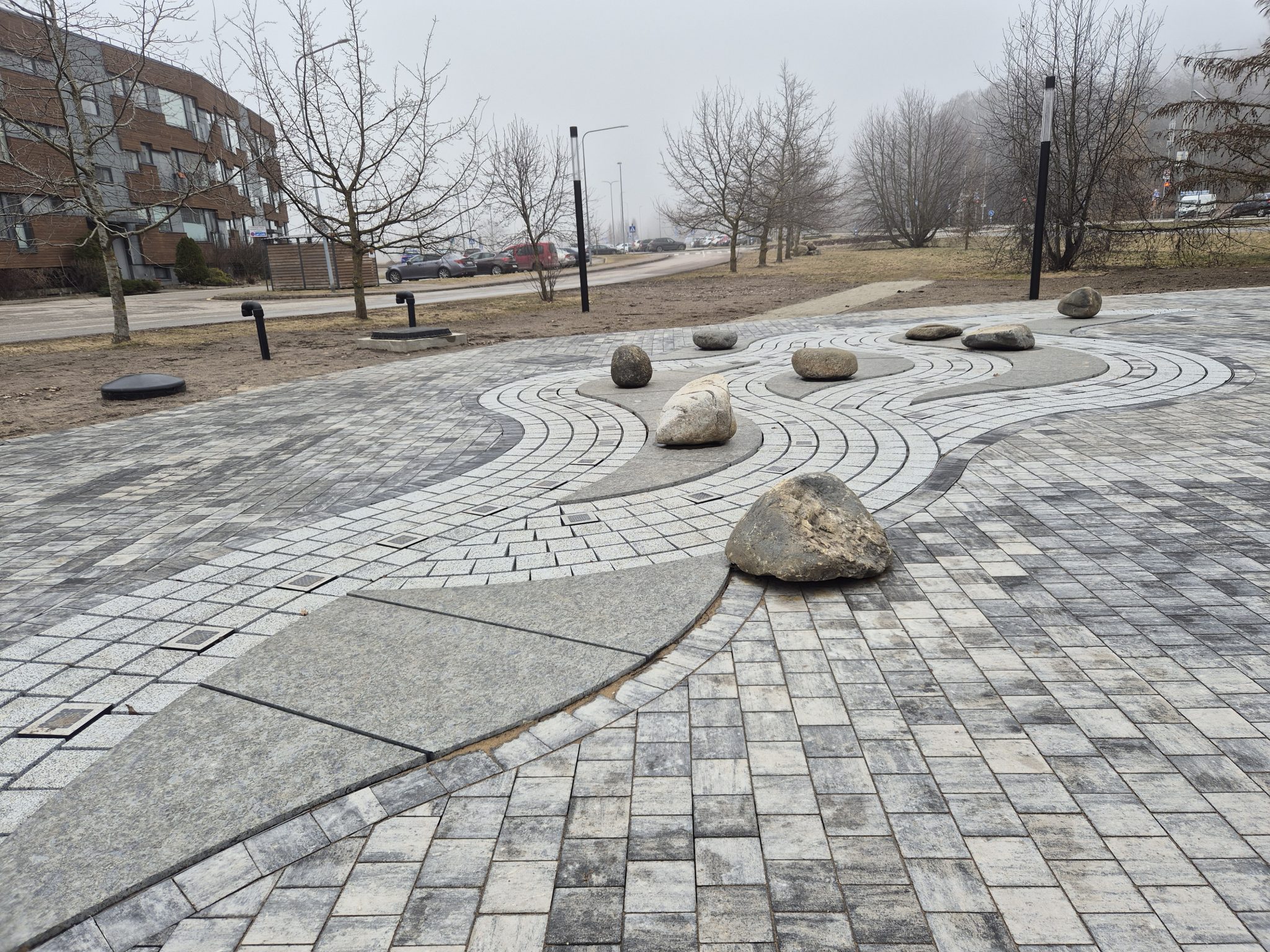Paved plaza with curved stone patterns and large individual rocks; a street, parked cars, and an apartment building in the background.