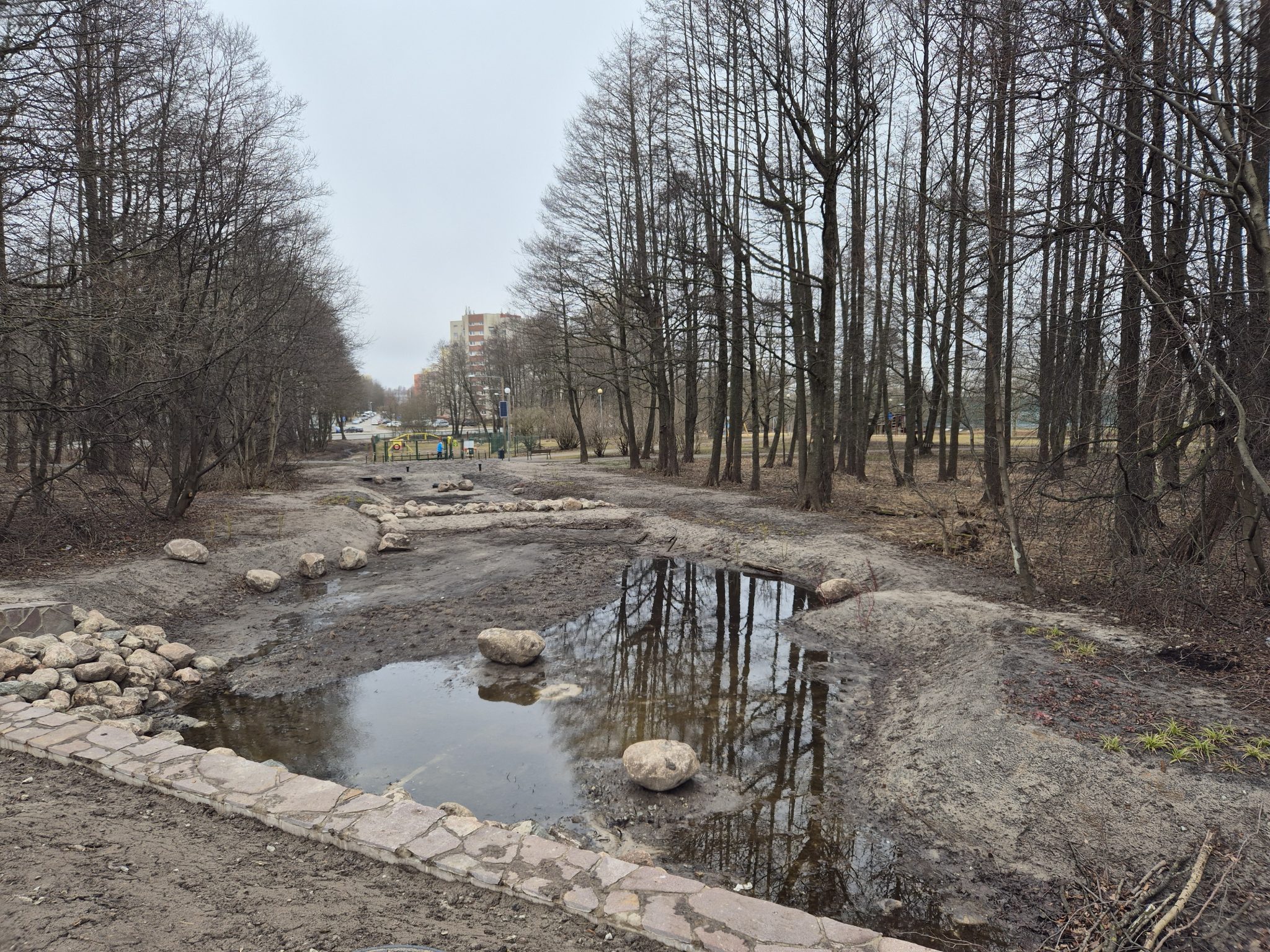 Puistomainen alue, jossa matala vesiallas ja kiviä, ympärillä puita ja taustalla asuinrakennuksia sekä leikkipuisto.