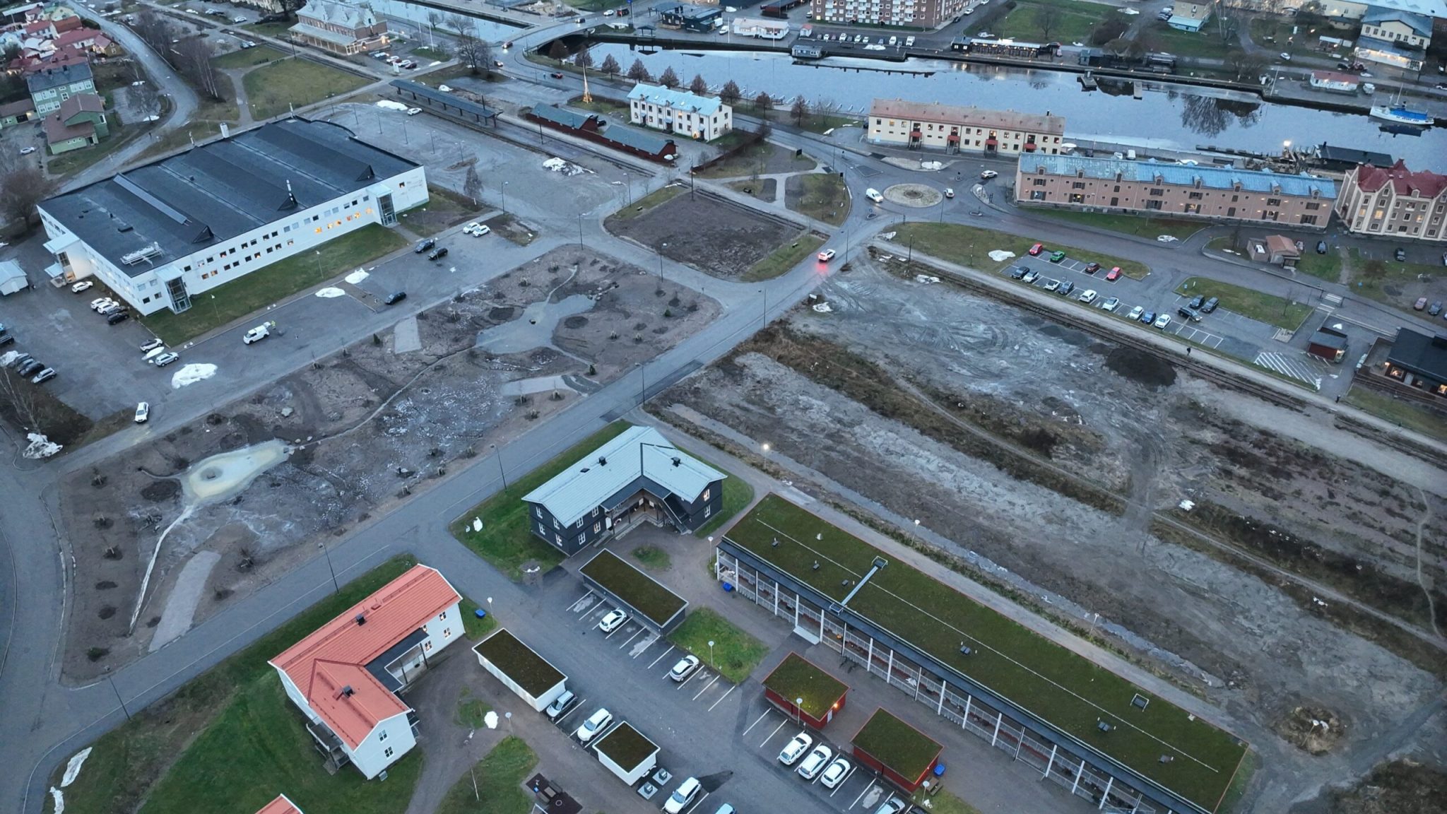 Ilmakuva kaupunkialueesta joen varrella: rakennuksia, pysäköintialueita ja osin rakentamatonta maata risteävien katujen ympärillä.