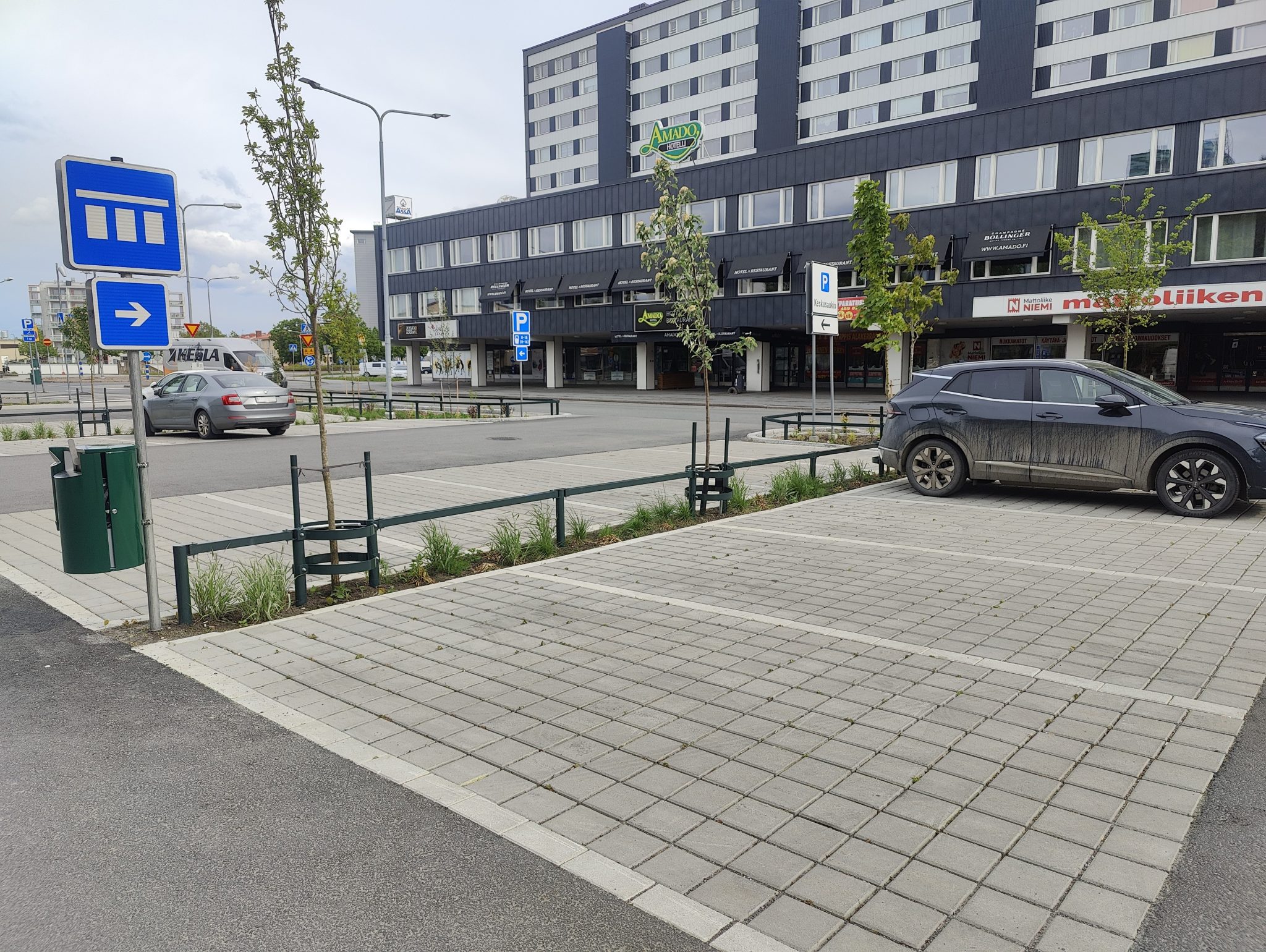 Urban street with a parked car, traffic signs, and an apartment building with ground-floor retail spaces; plantings and a sidewalk in front.