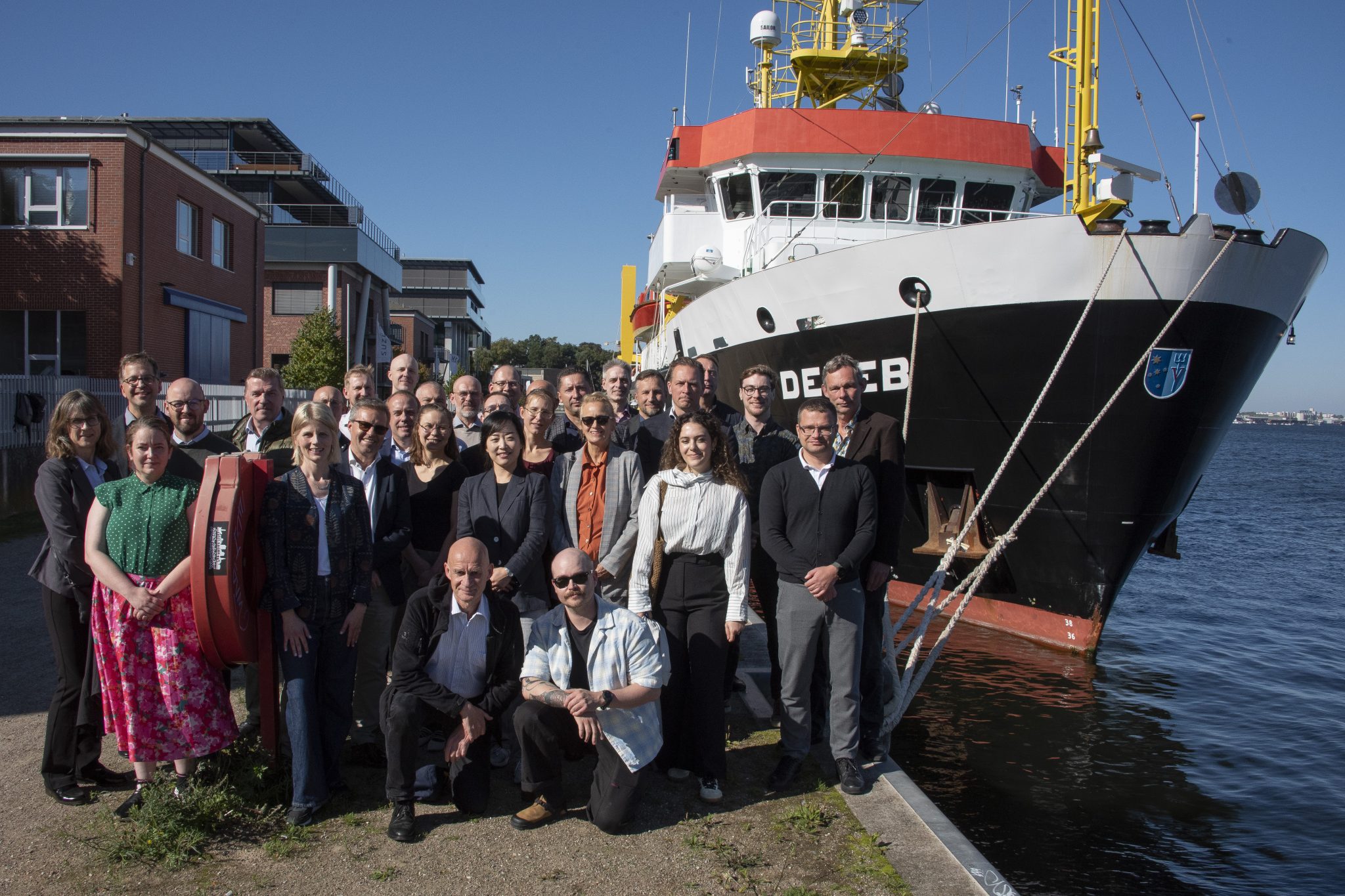 Group photo of event participants standing on a quay in front of the research vessel Deneb.