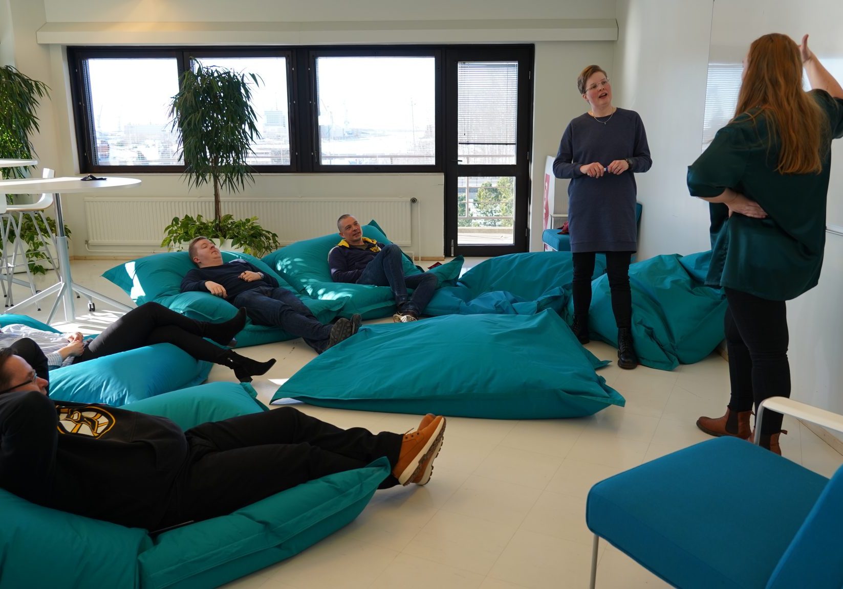 Two teachers and four persons sitting around on beanbag chairs listening.