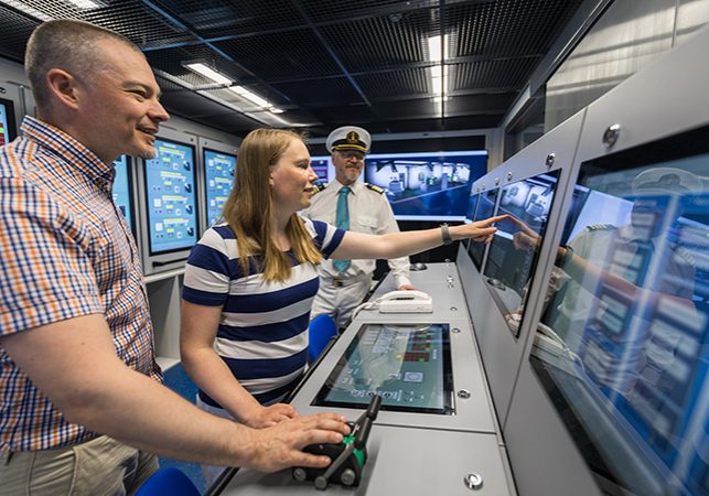 Kolme henkilöä seuraa merenkulunsimulaattorissa olevia monitoreja.