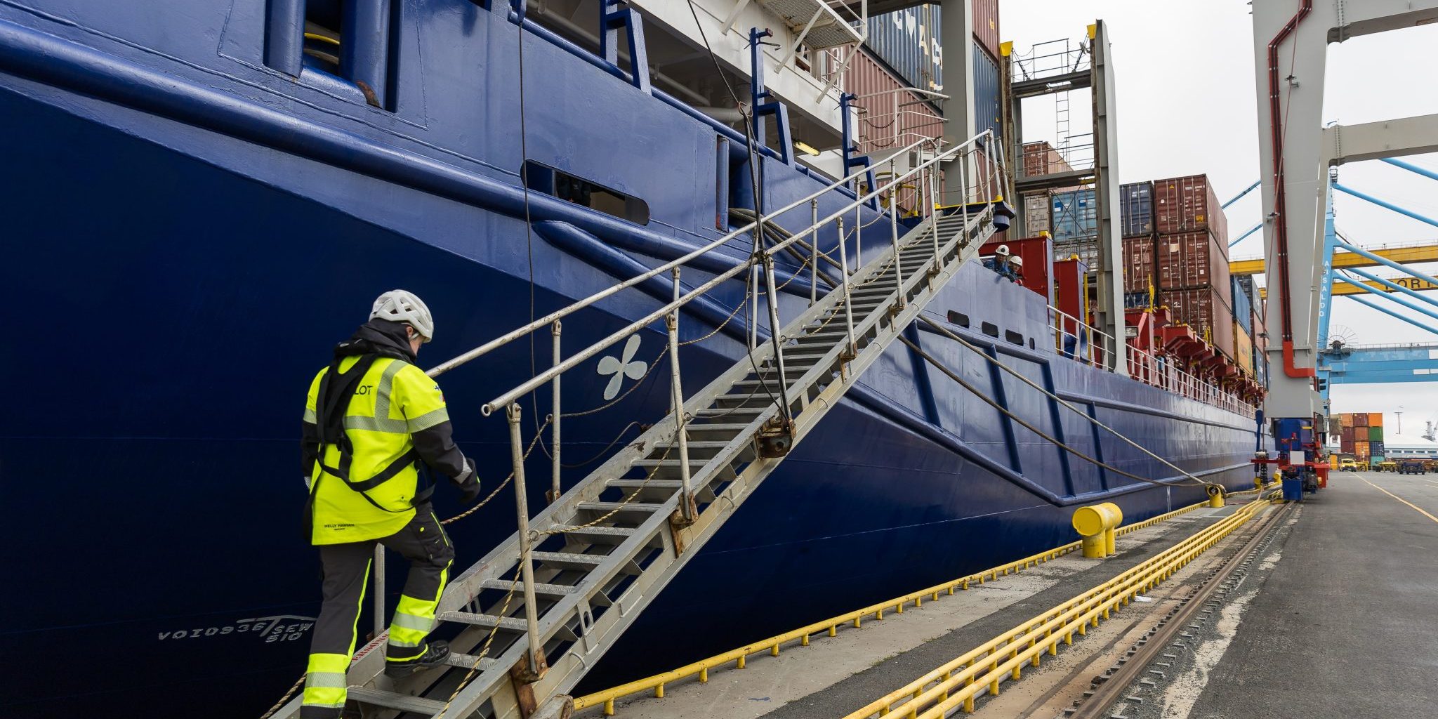 Person climbing stairs onto a cargo ship, wearing high-visibility clothing and a helmet.