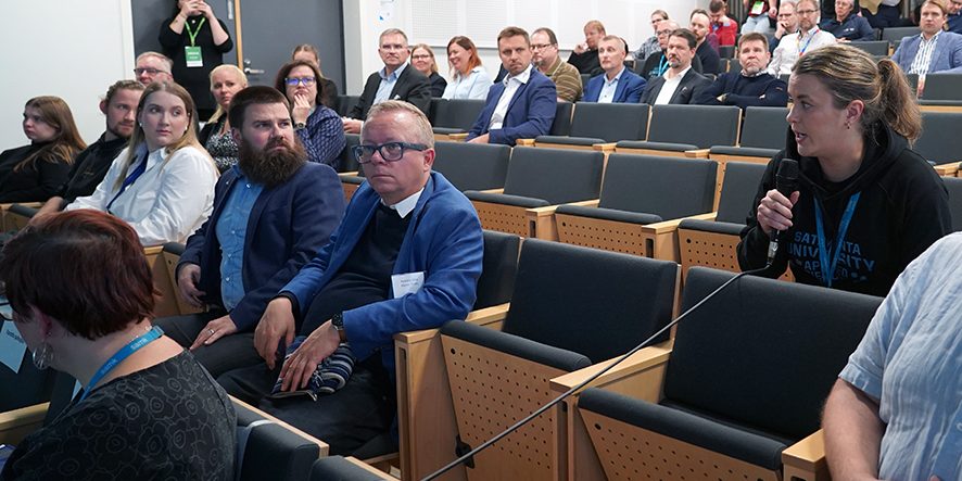 Tutkimusfoorumin yleisöä auditoriossa. Yleisöstä yksi henkilö puhuu mikrofoniin.